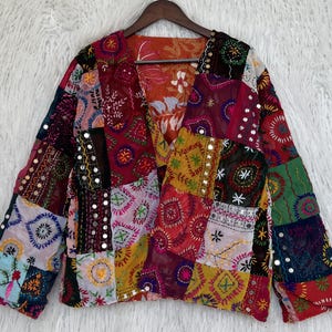 Peut inclure: Une veste à manches longues présentant un motif patchwork composé de divers tissus colorés. Le vêtement est orné de broderies complexes, de motifs circulaires et de petits miroirs réfléchissants sur toute la surface textile multicolore.