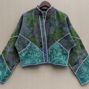 May include: A cropped jacket featuring a dark gray base with green floral embroidery. The garment has long sleeves, a mandarin collar, and a front opening accented with black and white striped trim. The lower section displays a teal green panel.