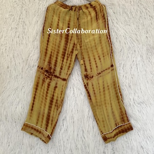 May include: Yellow and brown tie-dye pajama pants with white trim, hanging on a wooden hanger. The pants have a drawstring waist and a relaxed fit. The text "Sister Collaboration" is visible on the pants.