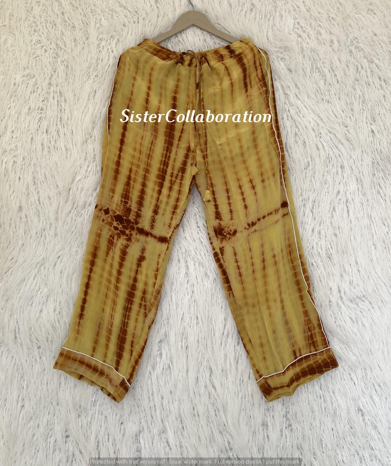 May include: Yellow and brown tie-dye pajama pants with white trim, hanging on a wooden hanger. The pants have a drawstring waist and a relaxed fit. The text "Sister Collaboration" is visible on the pants.