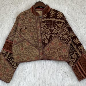 Op de afbeelding: Een kort jasje met een patchworkontwerp in diverse bruine, beige en donkerchocoladekleurige bloemen- en wervelpatronen. Het kledingstuk heeft een opstaande kraag, lange mouwen en een opening aan de voorzijde met zichtbare stiksels over de stof.