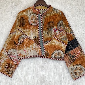 May include: A cropped jacket featuring a vibrant orange and brown quilted pattern with circular sun-like motifs. The garment has long sleeves, a mandarin collar, and a front opening trimmed with a thin, dark red and white checkered fabric border.