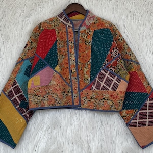 Op de afbeelding: Een patchworkjasje met een mandarijnkraag en lange mouwen. De jas heeft een verscheidenheid aan kleurrijke stoffen patches in tinten oranje, rood, blauwgroen en geel. De randen zijn afgewerkt met een blauwe bies.