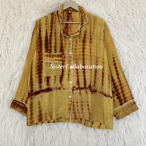 May include: A long-sleeved, button-down pajama top with a tie-dye pattern in shades of yellow and brown. The shirt has a chest pocket and white trim along the collar, cuffs, and pocket. The text "SisterCollaboration" is visible on the pocket.