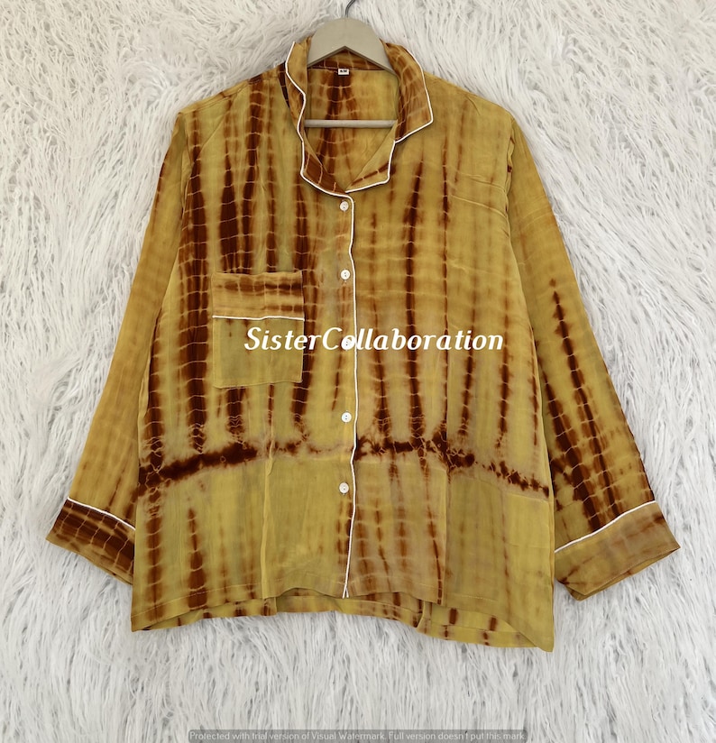 May include: A long-sleeved, button-down pajama top with a tie-dye pattern in shades of yellow and brown. The shirt has a chest pocket and white trim along the collar, cuffs, and pocket. The text "SisterCollaboration" is visible on the pocket.