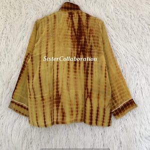 May include: A long-sleeved, tie-dye shirt in shades of yellow and brown. The shirt has a collared neckline and white trim on the cuffs and collar. The text "Sister Collaboration" is printed on the shirt.