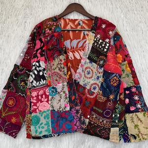 May include: A long-sleeve jacket featuring a patchwork design of various colourful fabrics with intricate floral embroidery and small reflective mirror accents. The garment has an open front and a visible interior lining with a brown leaf pattern.