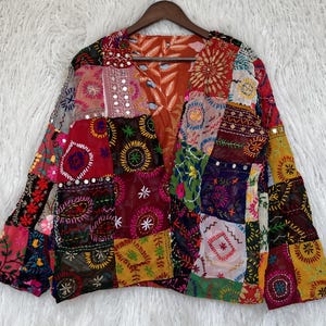 Peut inclure: Une veste à manches longues présentant un motif patchwork composé de divers tissus colorés. Le vêtement est orné de broderies complexes, de motifs circulaires et de petits miroirs réfléchissants sur tous les panneaux textiles multicolores.