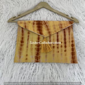 May include: A tie-dye clutch in shades of yellow and brown, with a gold tassel and the text "Sister Collaboration". The clutch is hanging on a wooden hanger against a white, fluffy background.