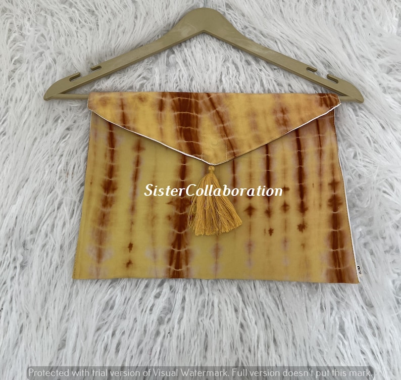 May include: A tie-dye clutch in shades of yellow and brown, with a gold tassel and the text "Sister Collaboration". The clutch is hanging on a wooden hanger against a white, fluffy background.