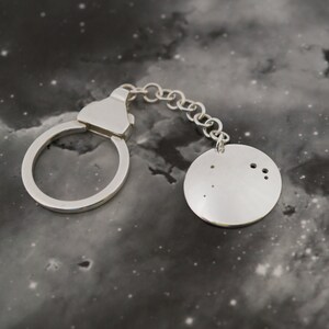 May include: Silver keychain with a round charm featuring a constellation design. The charm is attached to a silver ring and chain.