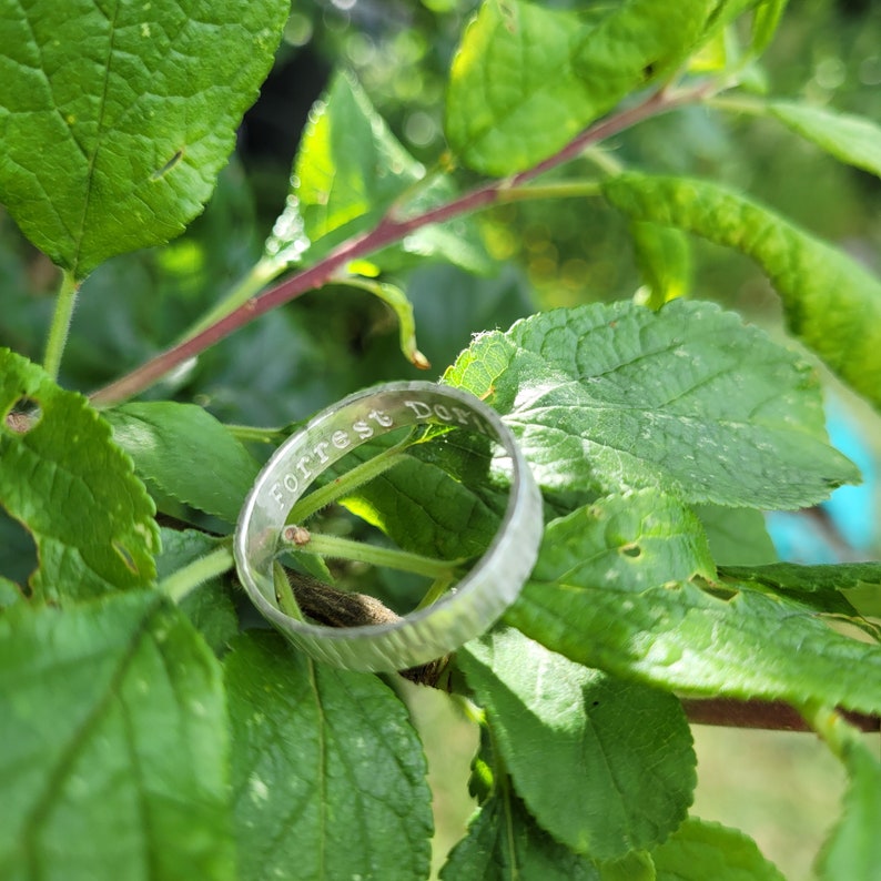 K&ouml;nnte beinhalten: Ein silberner Ring mit der Gravur "Forrest Dot" auf der Au&szlig;enseite des Bandes. Der Ring liegt auf einem gr&uuml;nen, belaubten Zweig.