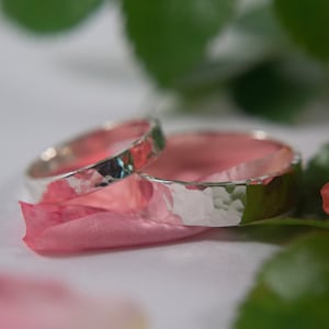 Op de afbeelding: Twee zilveren gehammerde trouwringen op een roze bloemblaadje.