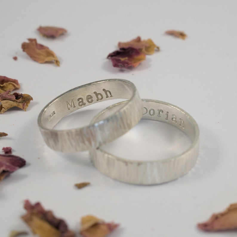 K&ouml;nnte beinhalten: Zwei silberne Ringe mit einer strukturierten Oberfl&auml;che. Die Ringe sind mit den Namen "Maebh" und "Dorian" graviert.