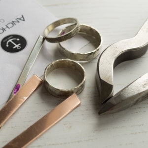 May include: Close-up of jewelry making tools and supplies, including three silver rings, two copper strips, and a metal ring-bending tool. The rings are hammered and have a textured finish.
