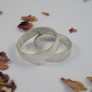 K&ouml;nnte beinhalten: Zwei silberne Ringe mit einer strukturierten Oberfl&auml;che. Ein Ring ist mit dem Datum "23.01.22" und der andere mit dem Namen "Maebl" graviert.