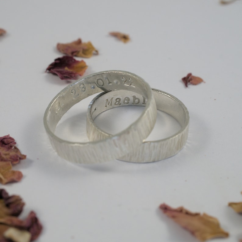 K&ouml;nnte beinhalten: Zwei silberne Ringe mit einer strukturierten Oberfl&auml;che. Ein Ring ist mit dem Datum "23.01.22" und der andere mit dem Namen "Maebl" graviert.