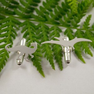 May include: A pair of silver cufflinks featuring a dinosaur and a pterodactyl design.