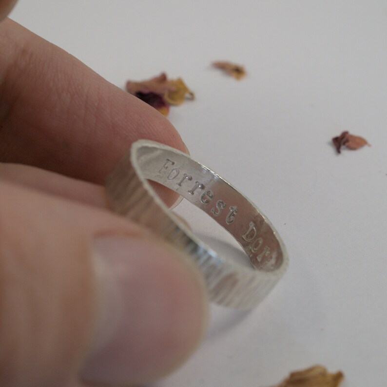 K&ouml;nnte beinhalten: Ein silberner Ring mit einer strukturierten Band und der Gravur "Forrest Dopp" auf der Innenseite.