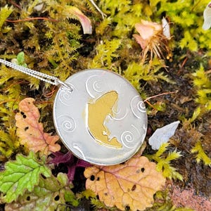 May include: A silver pendant necklace with a round charm featuring a gold silhouette of a fish. The charm has a textured background with swirling lines.