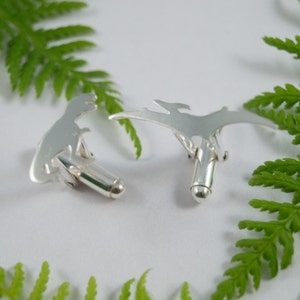 May include: A pair of silver cufflinks featuring a dinosaur and a pterodactyl silhouette. The cufflinks are on a white background with green fern leaves.