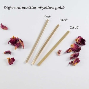 May include: Three gold bars of different purities: 9ct, 14ct, and 18ct. The bars are arranged in a row on a white surface with pink rose petals scattered around them.