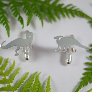 May include: A pair of silver dinosaur cufflinks. One cufflink features a silhouette of a Tyrannosaurus Rex, while the other cufflink features a silhouette of a Stegosaurus.