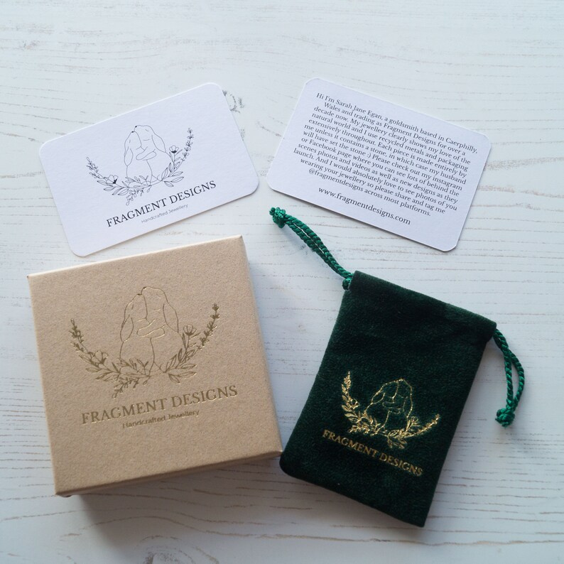K&ouml;nnte beinhalten: Eine kleine gr&uuml;ne Samt-Tasche mit einem goldenen Logo, auf dem "FRAGMENT DESIGNS" steht, und einer Zeichnung von zwei sich umarmenden Kaninchen. Die Tasche liegt auf einer wei&szlig;en Holzoberfl&auml;che. Neben der Tasche befindet sich eine kleine Karte mit dem gleichen Logo und Text. Die Karte enth&auml;lt auch eine Nachricht des K&uuml;nstlers.