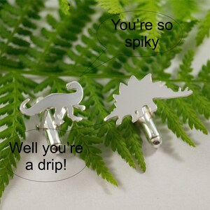 May include: A pair of silver dinosaur cufflinks. One cufflink is a stegosaurus with the text "You're so spiky" above it. The other cufflink is a brachiosaurus with the text "Well you're a drip!" below it.