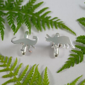 May include: A pair of silver cufflinks featuring a dinosaur and a triceratops silhouette. The cufflinks are displayed on a white background with green fern leaves.