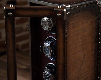 Puede incluir: Un enrollador de relojes de cuero marrón con forma rectangular y puerta abatible. El enrollador tiene un asa y capacidad para varios relojes. Los bordes están adornados con metal plateado. El enrollador está colocado frente a una pared de ladrillos.