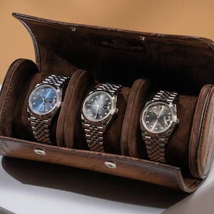 Może przedstawiać: Brązowe skórzane etui na zegarki z trzema srebrnymi zegarkami. Zegarki mają srebrne paski i ciemne tarcze. Etui jest otwarte, ukazując zegarki w brązowych przegródkach przypominających zamsz. Etui leży na białej powierzchni.