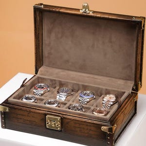 Peut inclure: Coffret à montres en bois marron ouvert, contenant huit montres. Le coffret est orné de ferrures en laiton et d'un intérieur gris. Les montres, aux designs variés, sont exposées, présentant une collection de garde-temps.