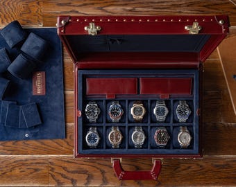 Puede incluir: Un estuche de reloj de cuero burdeos con interior azul marino, que contiene varios relojes de pulsera. El estuche está abierto, revelando múltiples compartimentos para los relojes. Un rollo de reloj y una tarjeta azules a juego están a la izquierda. Un cuaderno y gafas están a la derecha.