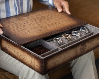 Puede incluir: Una caja de reloj de cuero marrón con un diseño de guión vintage en la tapa y los lados. El interior está forrado con tela gris y contiene varios relojes de pulsera. La caja está abierta, revelando los relojes.