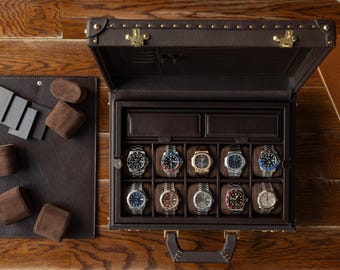 Puede incluir: Un estuche de reloj de cuero marrón con herrajes dorados, abierto para revelar diez relojes de pulsera. El estuche está sobre una superficie de madera, con almohadas de exhibición de relojes y una alfombrilla de cuero a la izquierda. Un cuaderno y gafas a la derecha.