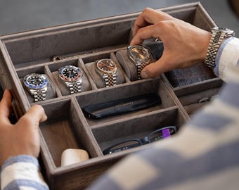 Puede incluir: Una caja de reloj de madera con interior de terciopelo gris, que contiene varios relojes de lujo con correas plateadas y doradas. Los relojes tienen esferas negras, azules y marrones. Una mano alcanza uno de los relojes.