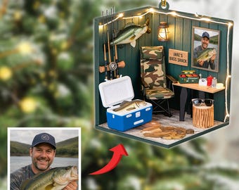 May include: A decorative ornament depicting a fishing scene. The ornament features a miniature fishing shack with a camouflage chair, a cooler with a fish, and a photo of a man holding a fish. The text "Jake's Bass Shack" is visible.