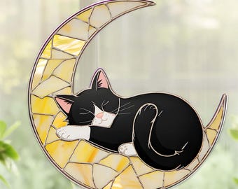 Puede incluir: Una decoración de media luna de vidriera con un gato blanco y negro durmiendo. La luna está hecha de vidrio de mosaico amarillo y blanco, y el gato está acurrucado sobre ella. Se adjunta una cadena negra para colgar.
