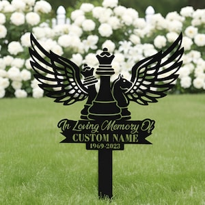 May include: Black metal memorial garden stake featuring chess pieces with angel wings. The stake reads "In Loving Memory Of" and includes space for a custom name and dates. The background shows white flowers and green grass.