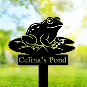 Puede incluir: Estaca de jardín de metal negro con una rana sobre una hoja de nenúfar. La rana tiene detalles blancos y está colocada sobre un letrero rectangular que dice "Celina's Pond". La estaca está colocada en hierba verde.
