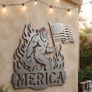 Peut inclure: Une décoration murale en métal représentant une silhouette découpée de Bigfoot portant un bandana, tenant un drapeau américain et entourée de flammes. Le mot MERICA est gravé en grandes lettres capitales au bas de cette plaque métallique.