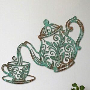 Puede incluir: Una pieza de arte mural de metal que muestra una tetera vertiendo líquido en una taza de té. El diseño tiene un acabado de pátina desgastada en tonos turquesa y cobre con intrincados patrones de hojas en espiral recortados.