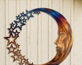 Könnte beinhalten: Ein Wandkunstobjekt in Form eines Halbmondes aus Metall mit warmem Bronzefinish. Der Mond zeigt ein menschliches Gesicht im Profil und ist von einer Reihe offener Sterne umgeben, deren Farbe sich von Bronze zu tiefem Blau hin verändert.