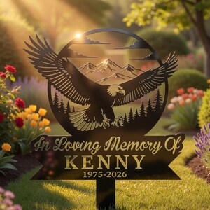 May include: Black metal memorial garden stake featuring a soaring eagle design within a circular frame, mountains, and trees. The text "In Loving Memory Of KENNY 1975-2026" is displayed on a banner below the eagle. The stake is placed in a garden.