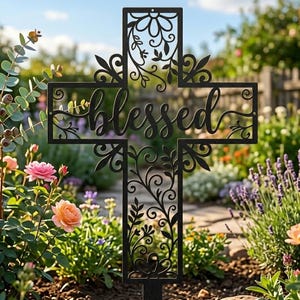 Puede incluir: Un estaca de jardín de metal negro con forma de cruz y patrones florales y de enredaderas cortados con láser. La palabra blessed está escrita en letra cursiva en el centro de la cruz. La estaca está diseñada para insertarse en el suelo.