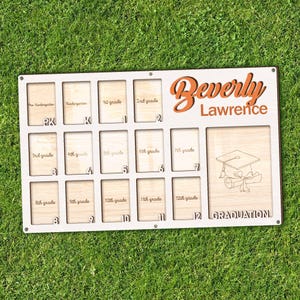 May include: A personalized wooden graduation display board with spaces for photos from pre-kindergarten through 12th grade. The board is white with the name "Beverly Lawrence" in orange script. A graduation cap and scroll are etched on the right.