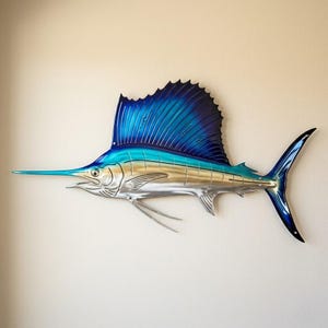 Op de afbeelding: Metalen wandkunst van een zeilvis. De sculptuur heeft een blauw-zilveren kleurenschema, met een grote rugvin en een lange, puntige snavel. De vis is bevestigd aan een neutraal gekleurde muur.