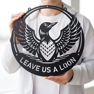 Może przedstawiać: Czarna metalowa grafika ścienna przedstawiająca nurka z rozłożonymi skrzydłami, umieszczona w okrągłej ramie. Tekst "LEAVE US A LOON" jest wyświetlany na dole. Projekt jest wycięty, tworząc efekt sylwetki.