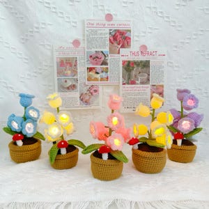 May include: Five crochet flower arrangements in small brown pots. Each arrangement features colorful flowers, green leaves, and a red mushroom. The flowers are blue, yellow, pink, and purple. The flowers have lights inside.